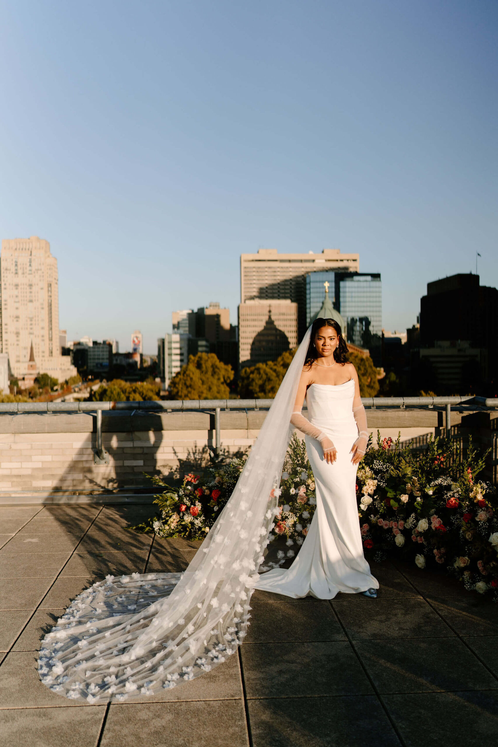 bride standing in direct sun on the roof of the Franklin Institute, staring into the camera with her veil spread out behind her, the Philadelphia skyline in the distance