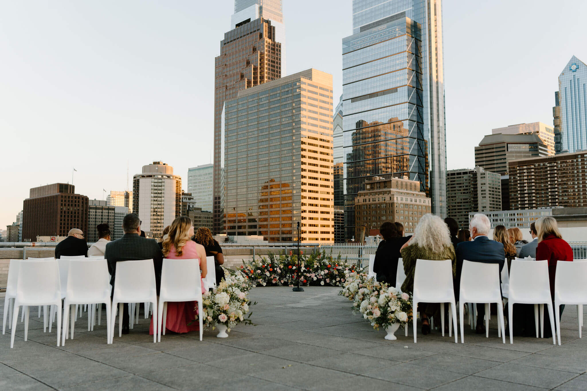 guests seated in white chairs on the roof, facing the Philadelphia skyline at sunset with rows of flowers lining the aisle and a floral arrangement at the altar of a franklin institute wedding ceremony