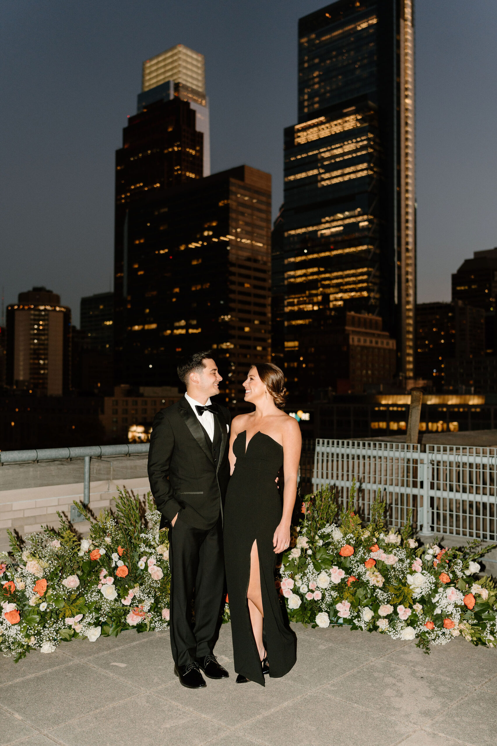 a couple, both wearing black, smiling at each with the lights of the Philadelphia skyline at dusk behind them