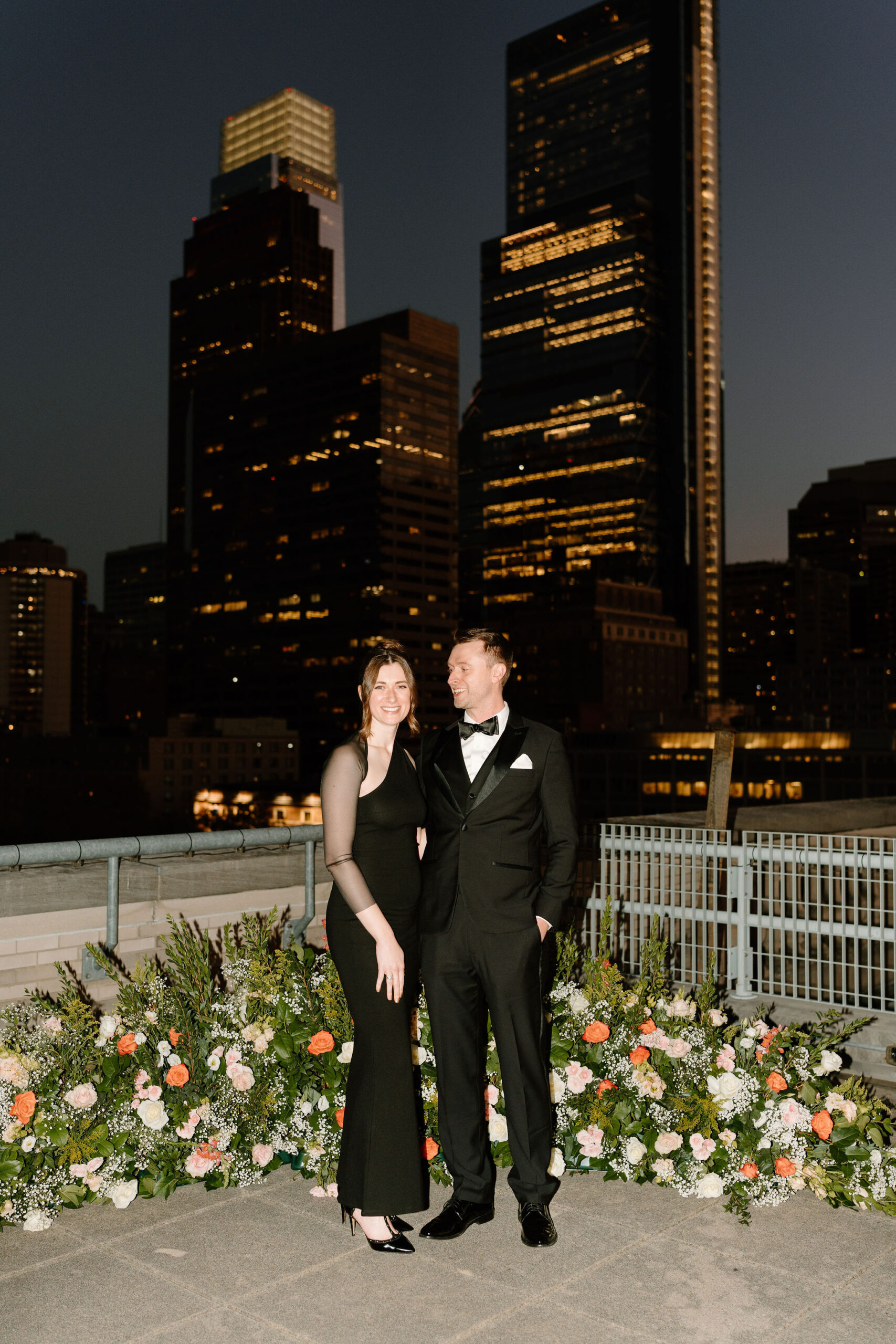 a couple, both wearing black, smiling at each with the lights of the Philadelphia skyline at dusk behind them