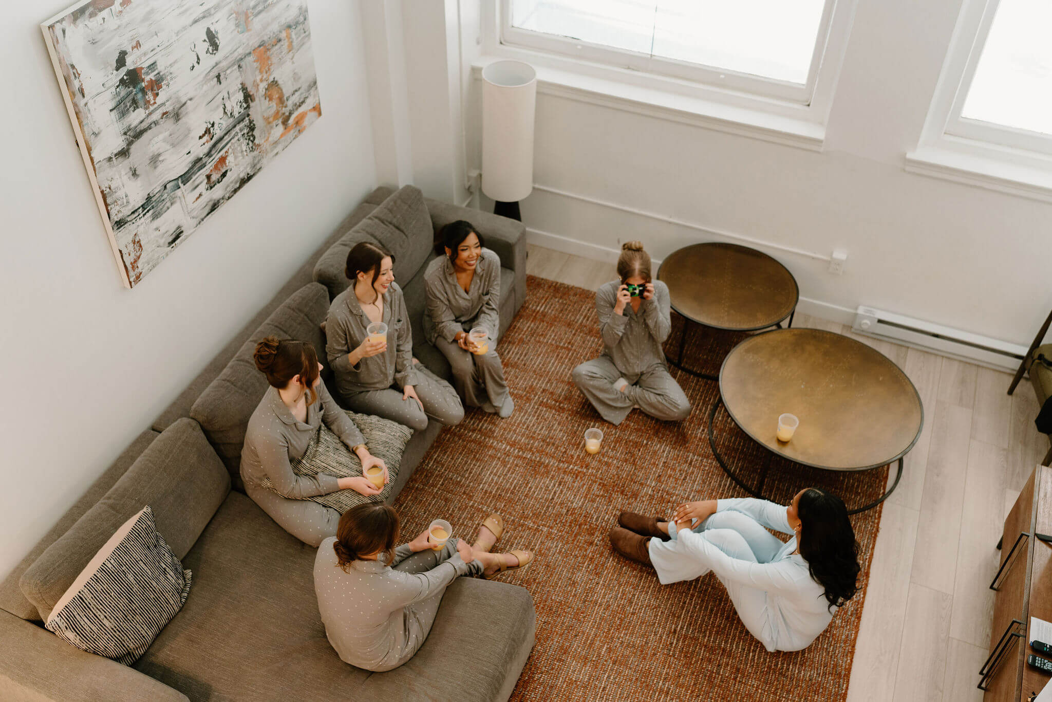 aerial image of bride (blue pajamas) sitting on the floor talking with five bridesmaids, also seated, laughing while one takes a disposable camera image 