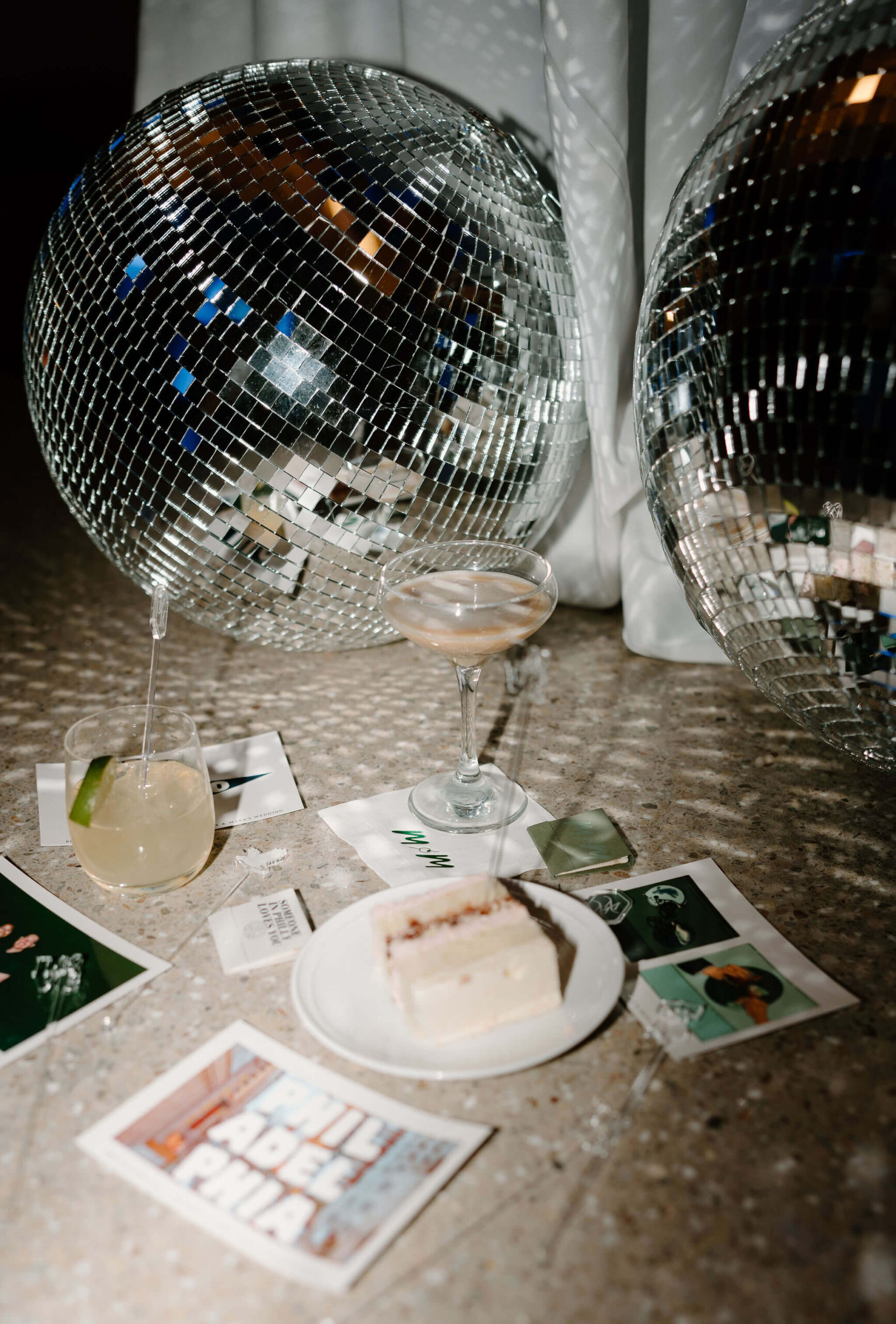 late-night flat lay at a wedding reception, featuring an espresso martini, a mule, a slice of wedding cake, post cards, a matchbox, and disco balls