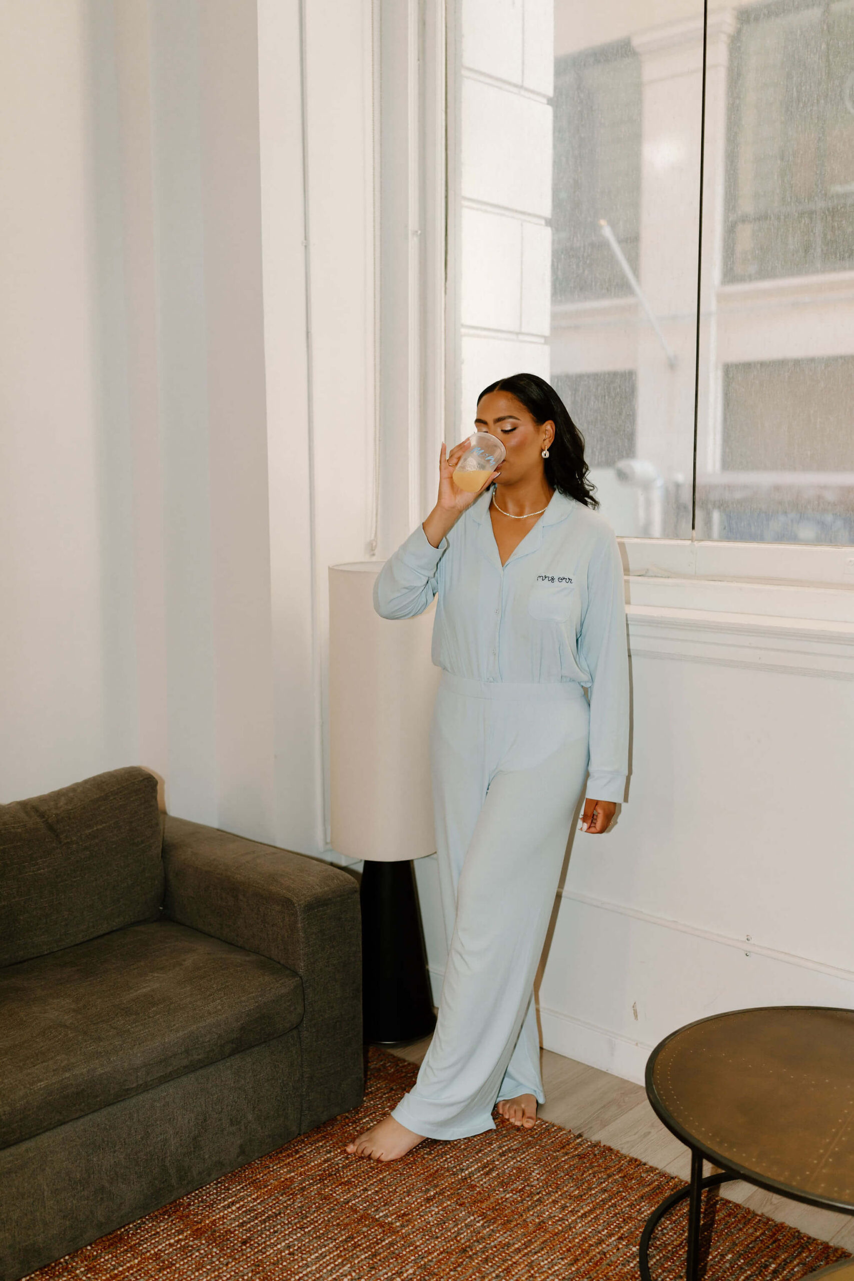 bride in custom blue Skimms pajamas with her name embroidered on the pocket, sipping a drink while getting ready on her wedding day