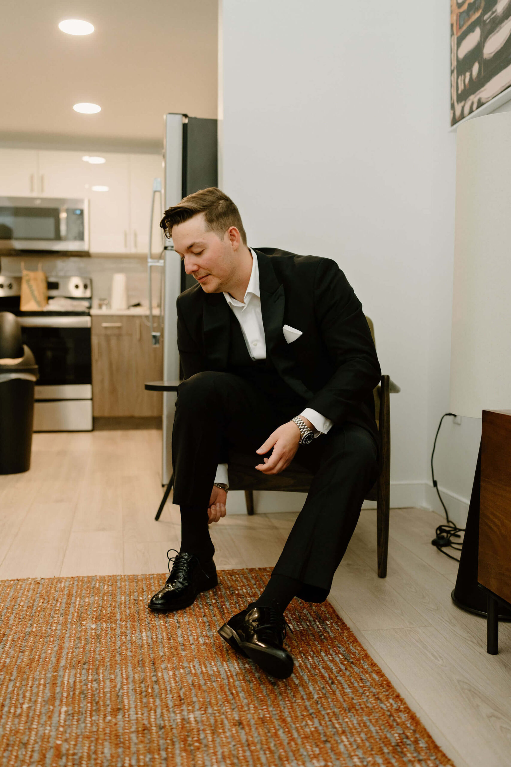 groom in black suit, seated in a chair and putting on his dress shoes