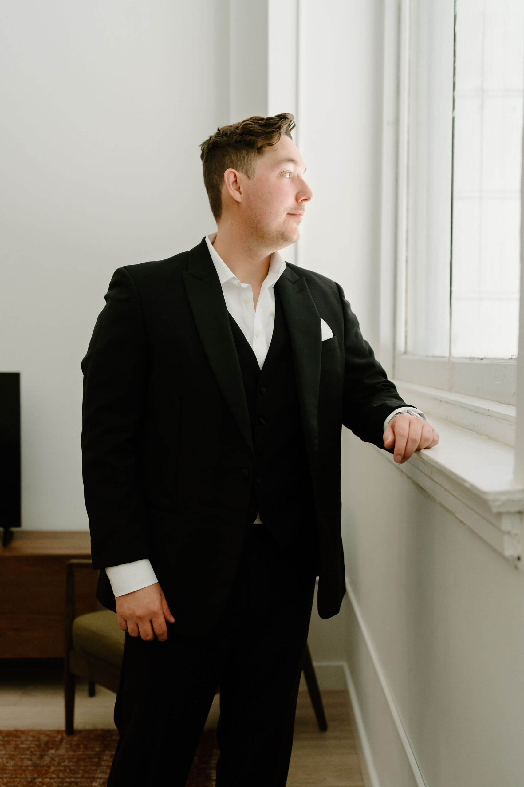 groom in black suit leaning on window frame and looking out window