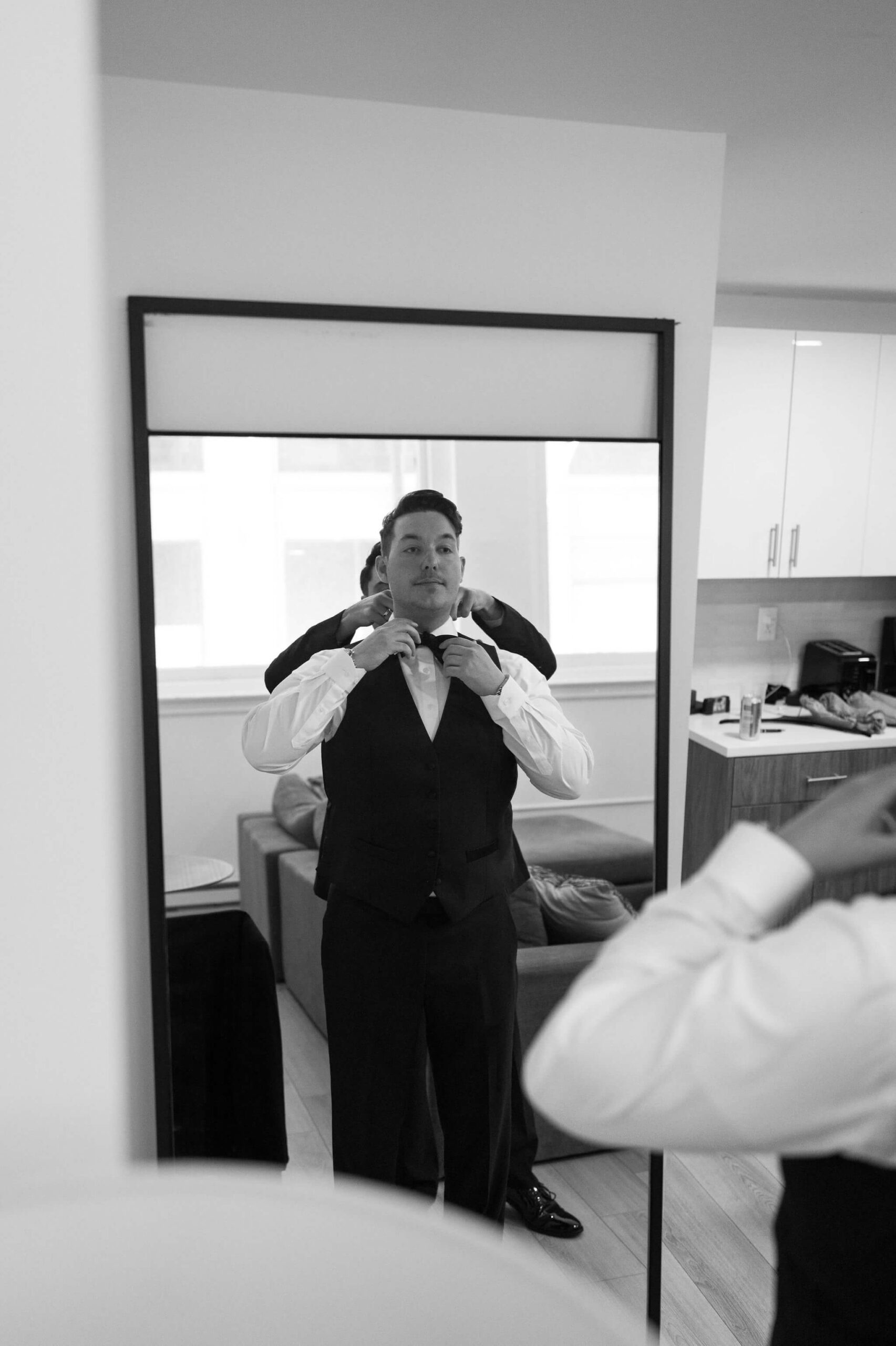 black and white image of groom in a mirror, being helped by a groomsman to put on his tie