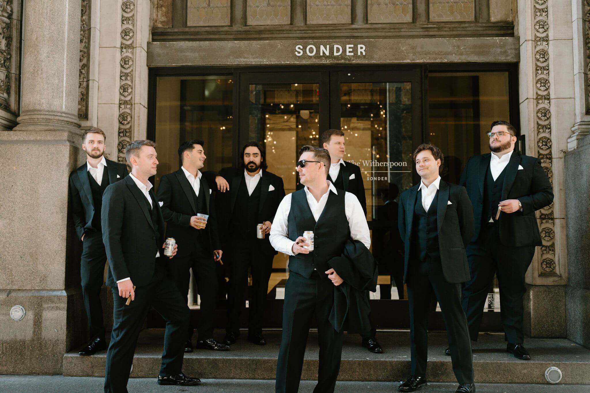 groom and groomsmen in black suits standing on the steps in front of a building with a sign that reads "Sonder", looking at each other and talking
