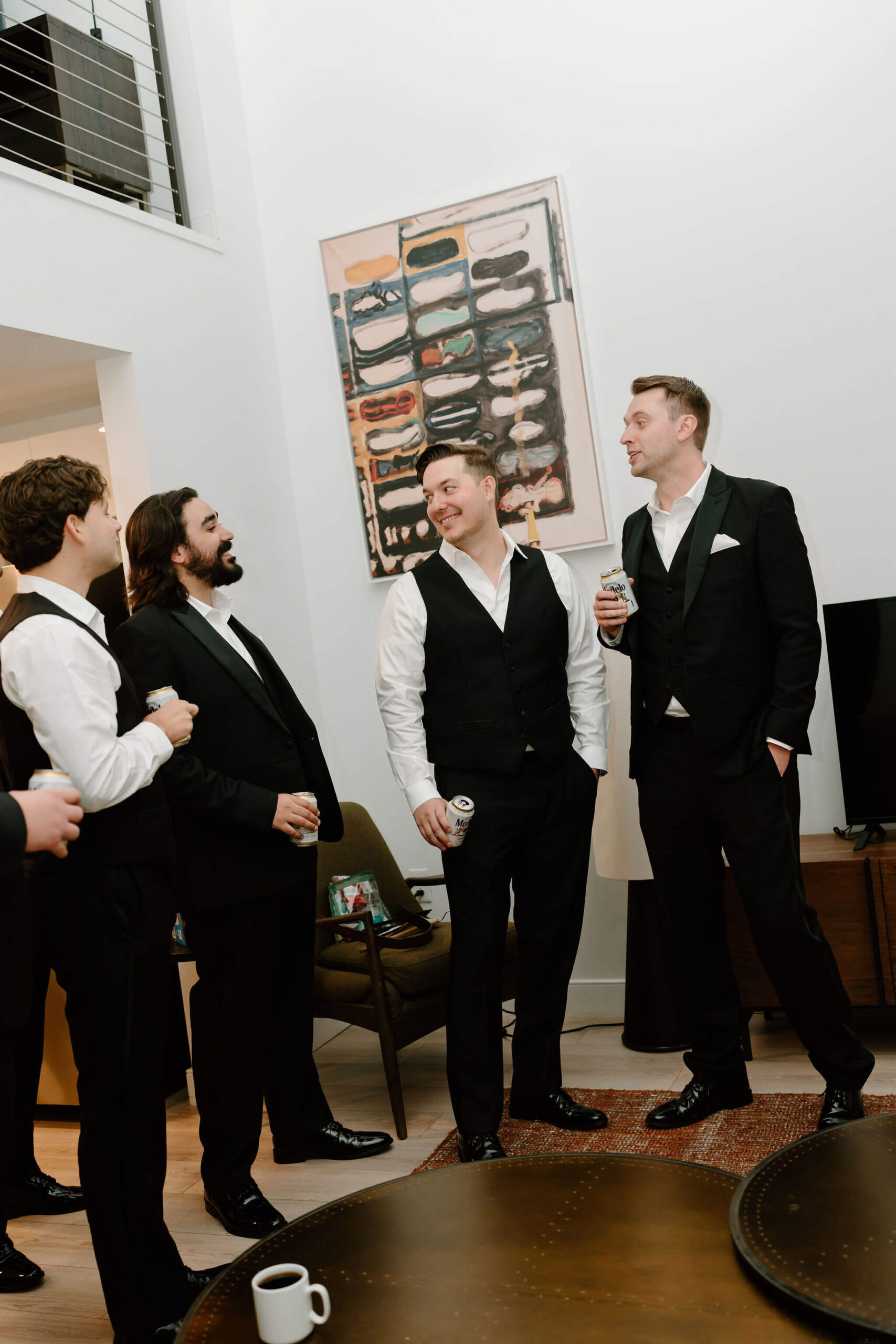 groom in black suit, holding a beer, surrounded by other groomsmen in black suits smiling and talking