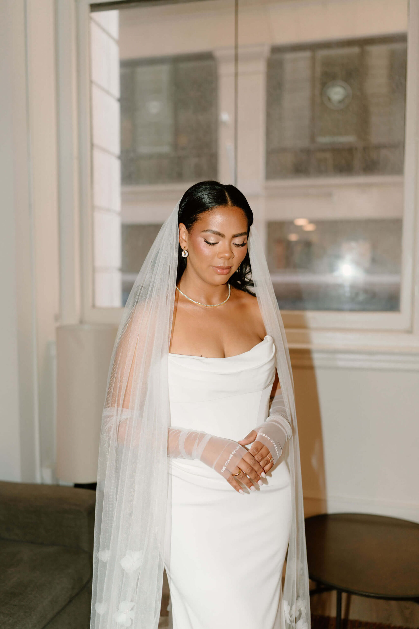 bride in her wedding gown, eyes cast down, with her hands gently held in front of her. She wears a long veil and gloves that read "you're my golden hour / the color of my sky"