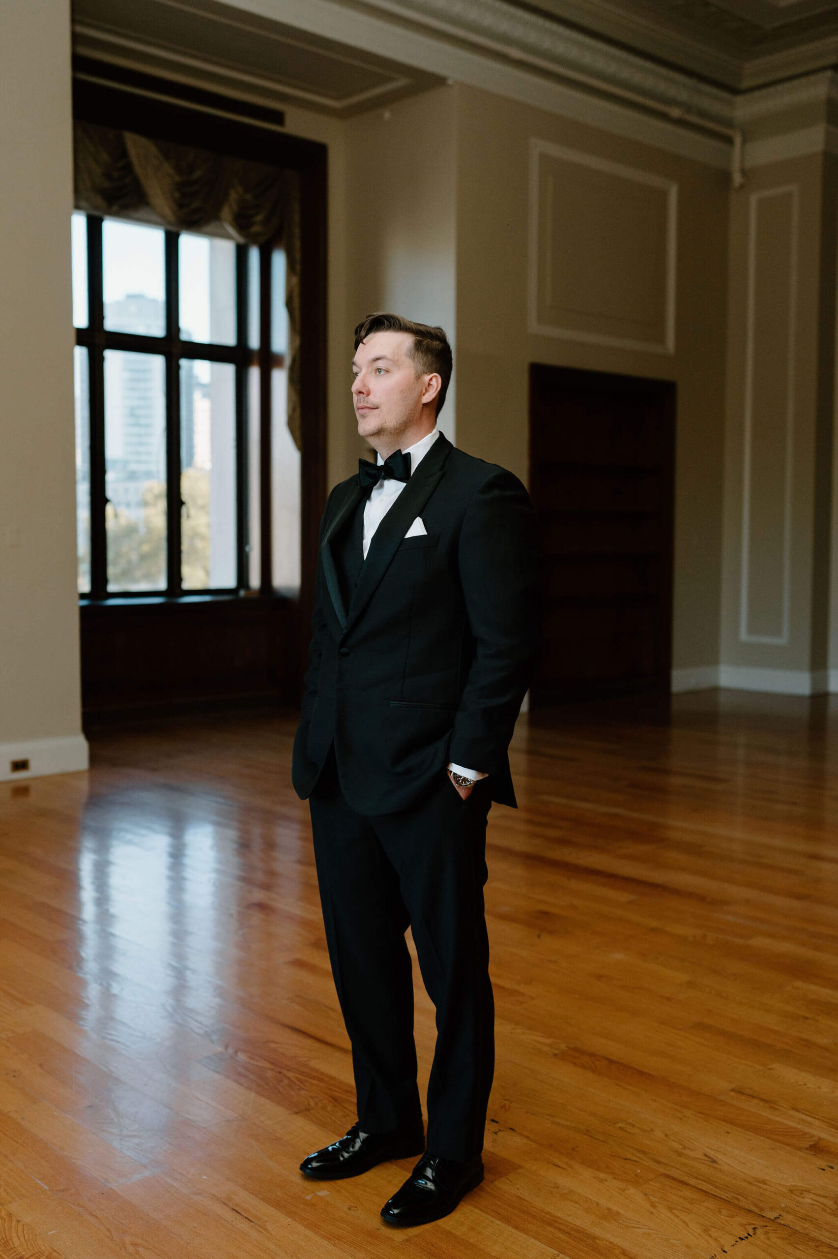 groom in a black suit with a black bowtie standing in a large empty room with a large window behind him, staring off camera at their franklin institute wedding