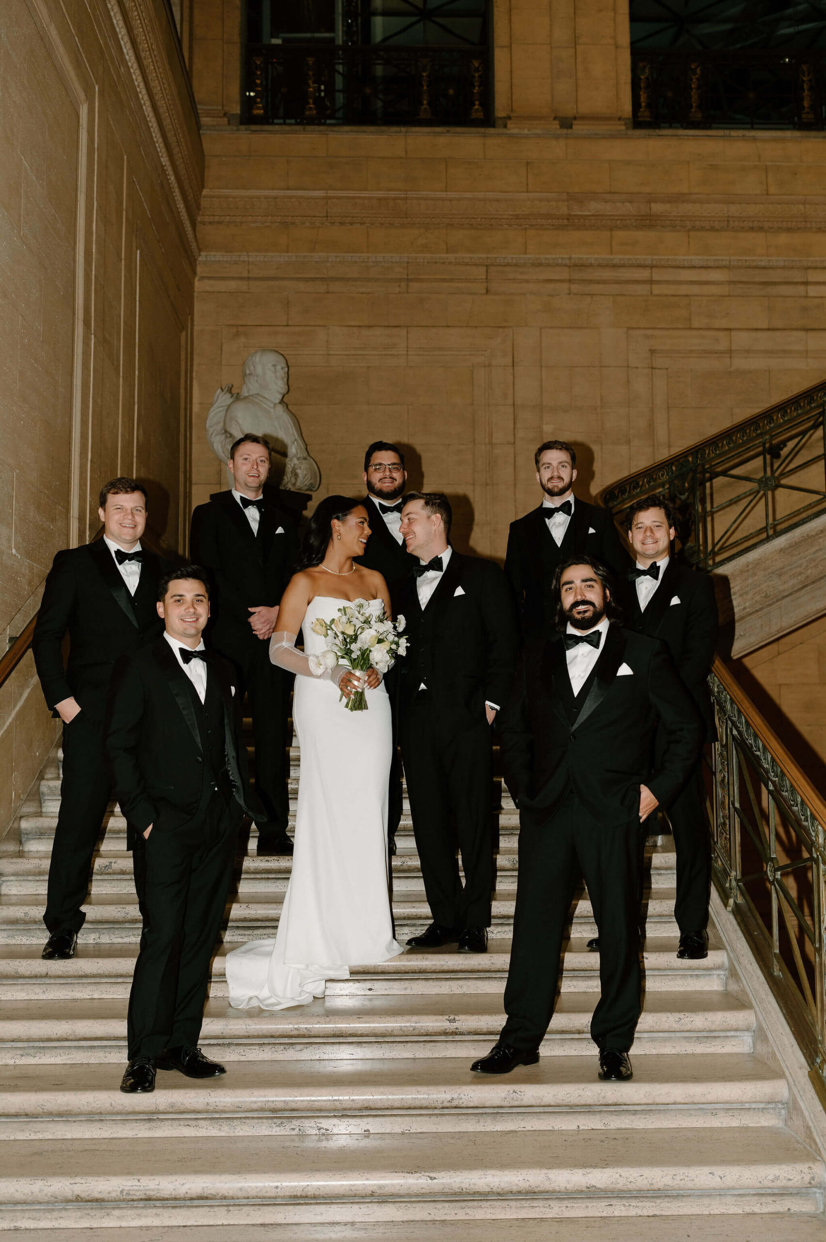 bride, groom, and groomsmen, all in formalwear, smiling at each other on a marble staircase