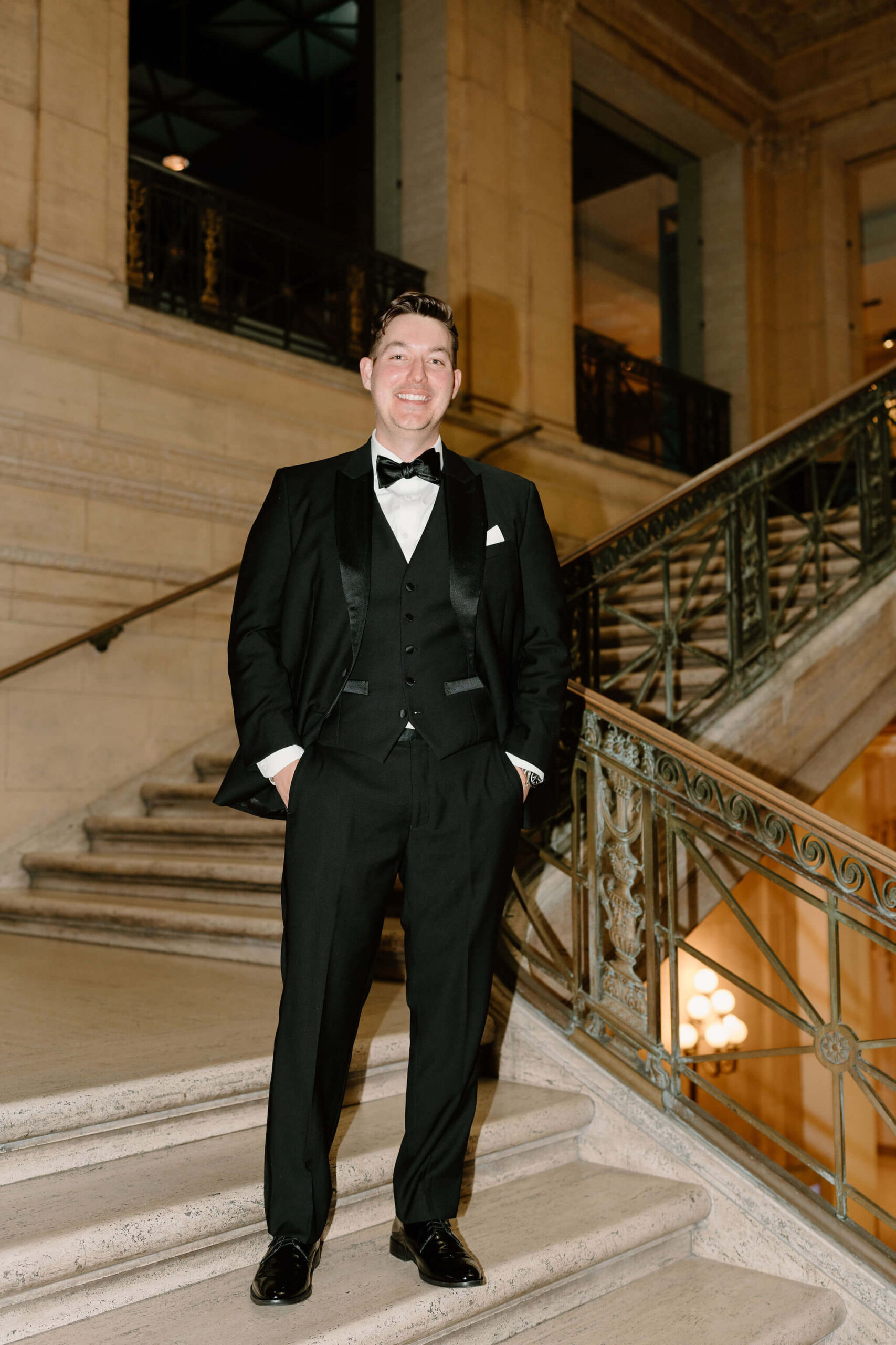 groom in a black tuxedo and black bowtie smiling widely at the camera on his wedding day, hands in his pockets, while standing on a marble staircase