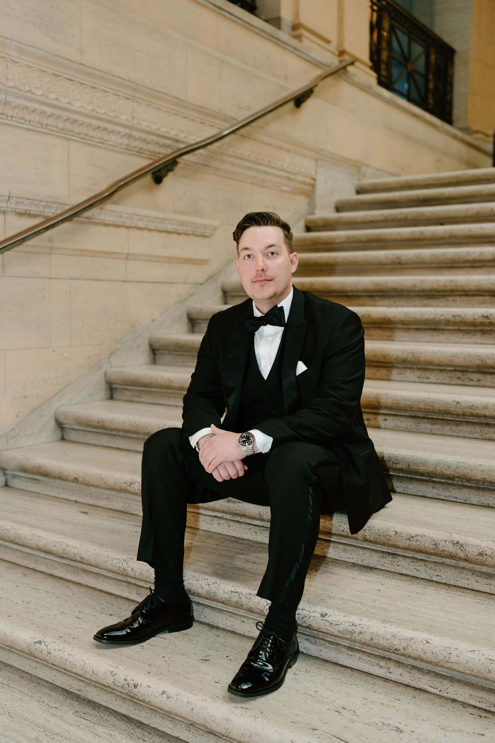 groom sitting on a marble staircase, hands clasped in front of his knees as he looks at the camera without smiling