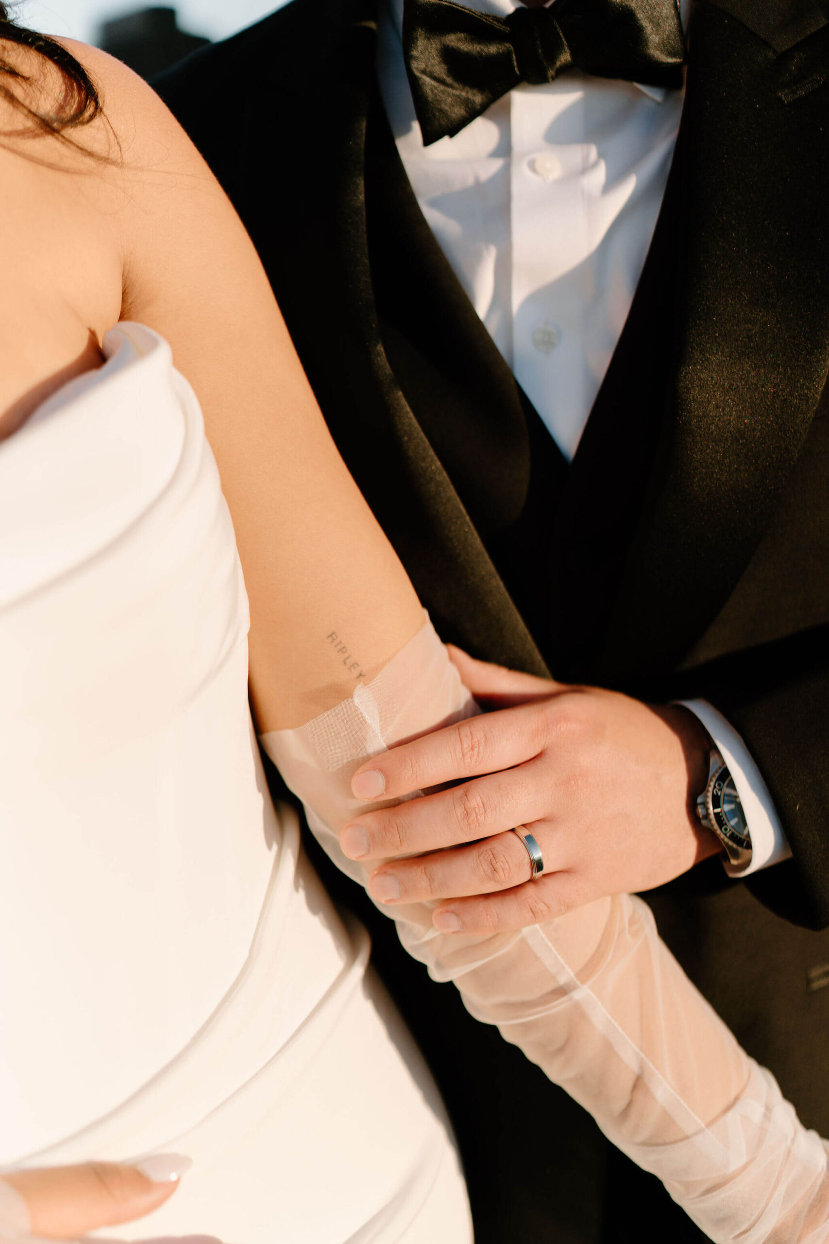 close-up of the groom's hand on the bride's arm, featuring his wedding ring and a tattoo just above her sheer gloves that reads "RIPLEY"
