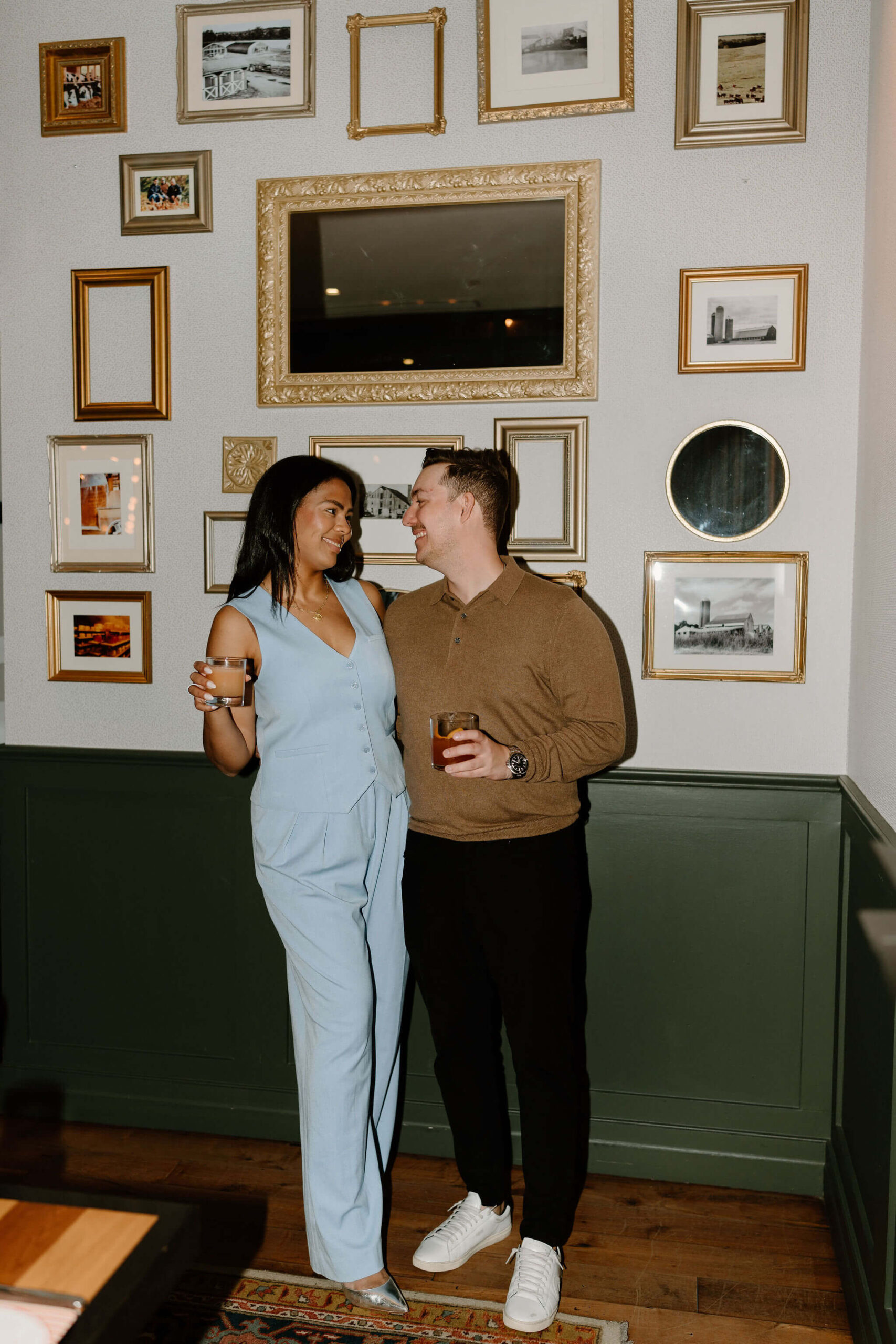 bride (baby blue jumpsuit, cocktail) smiling at groom (brown shirt, black pants, cocktail) against a wall of gold picture frames at their rehearsal dinner