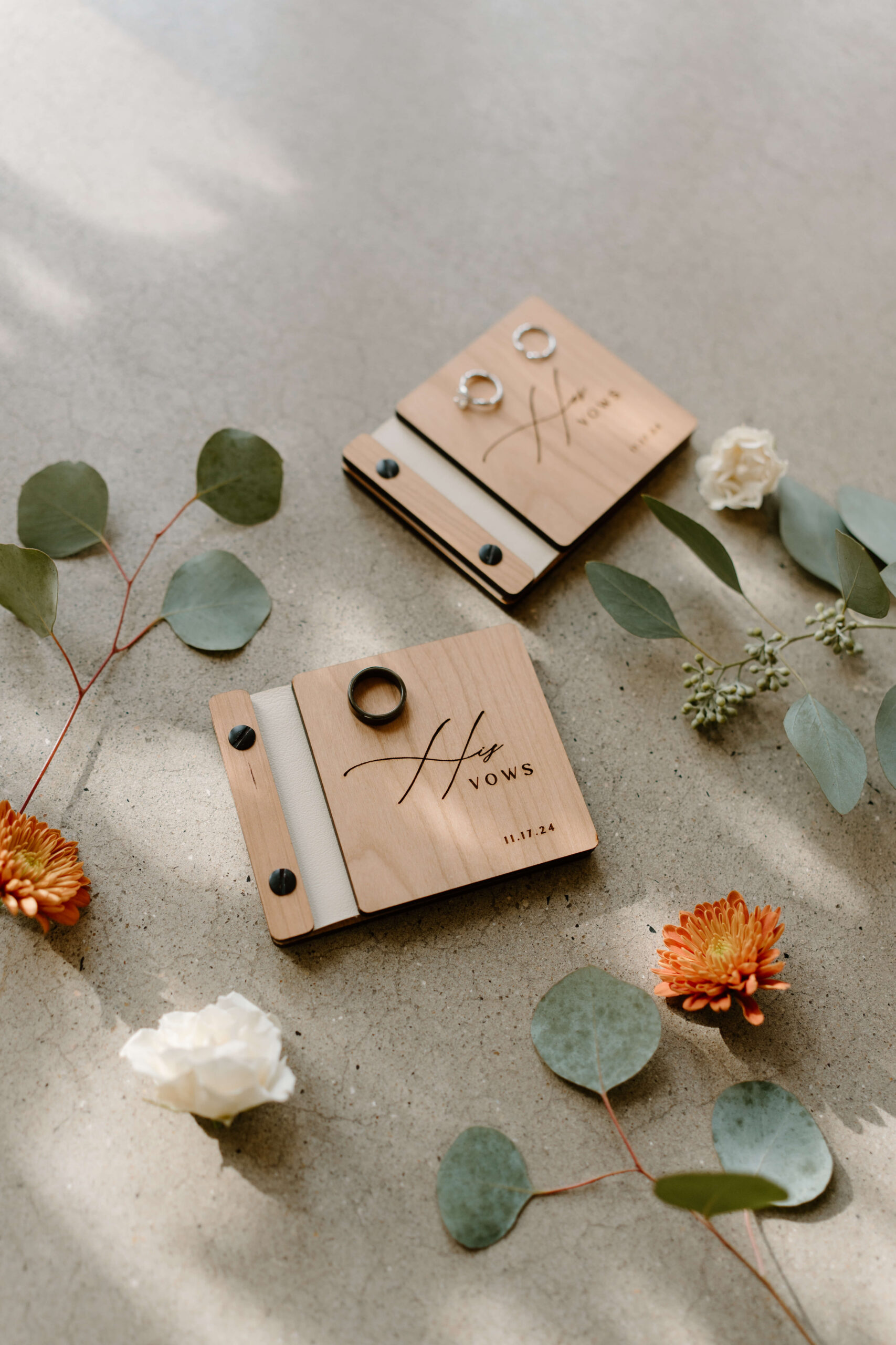 "his" and "hers" vow books, with two wedding bands and and engagement ring, surrounded by florals and eucalyptus branches