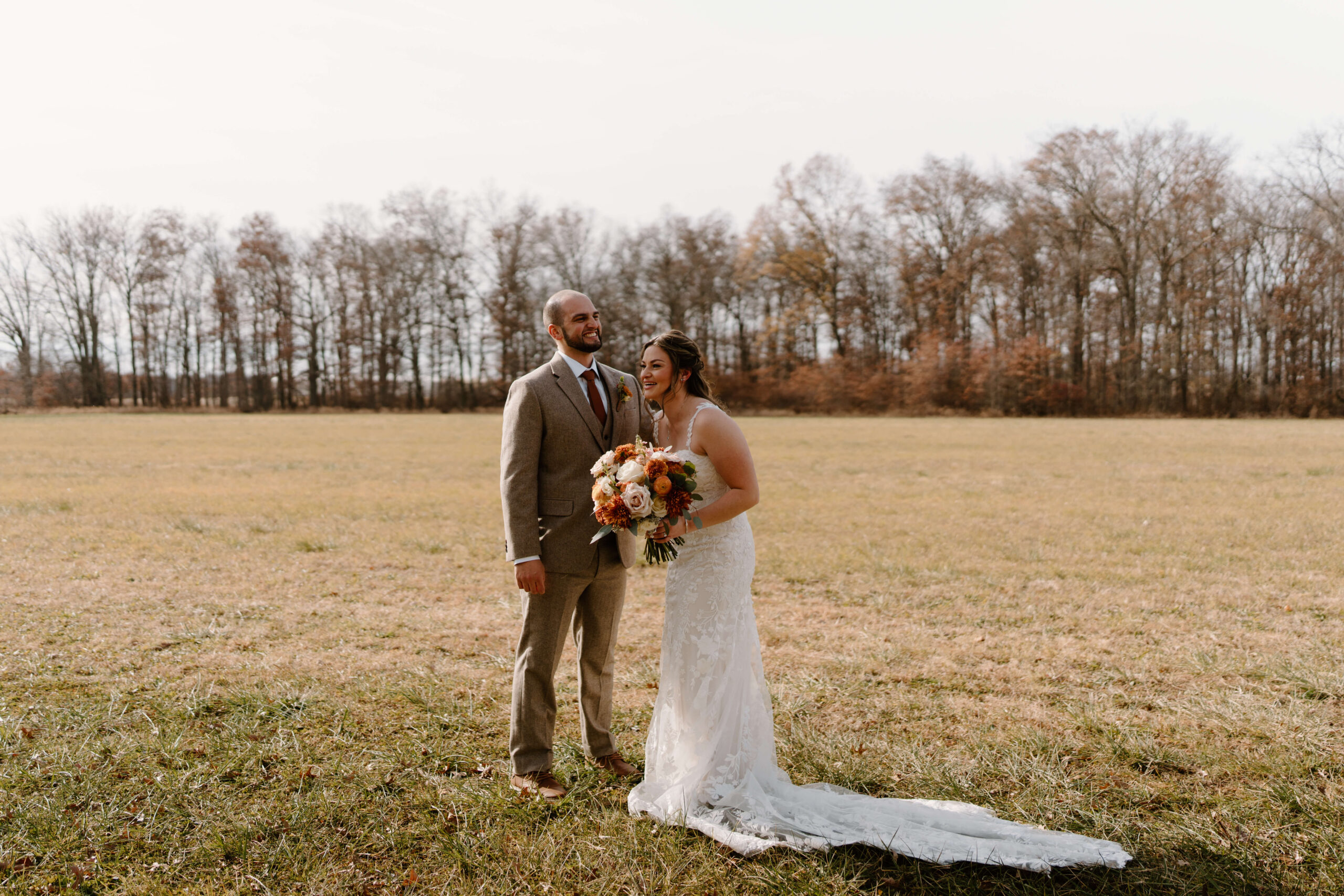bride (white wedding dress, holding flowers) and groom (brown tweed suit) laughing