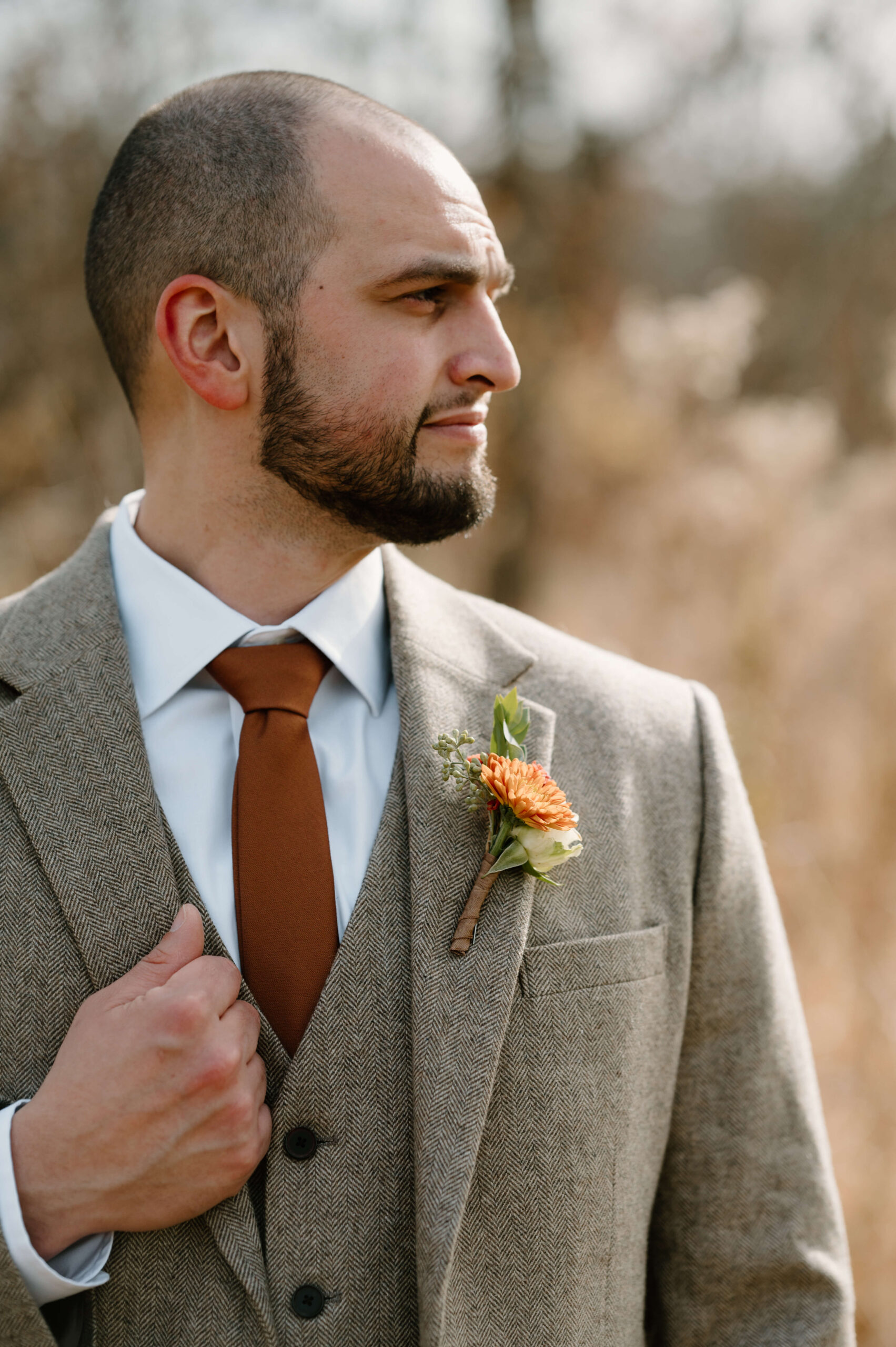 groom looking off-camera, holding the lapel of his tweed suit and wearing an orange-flowered boutonniere and rust-colored tie