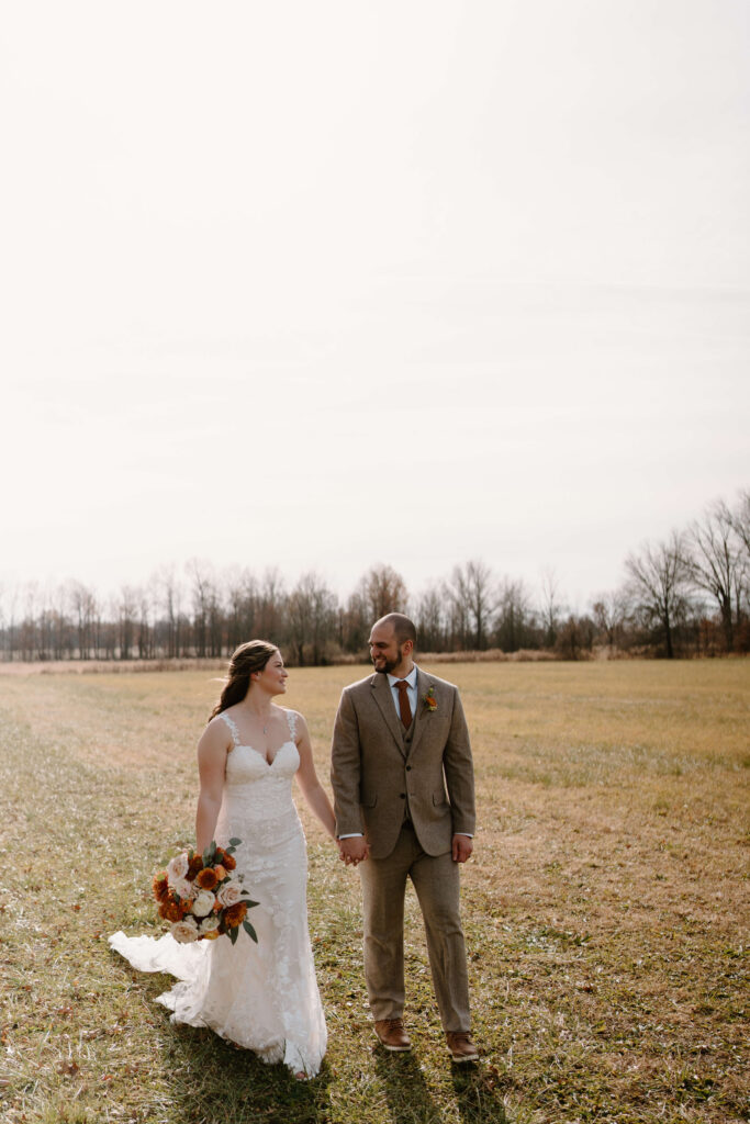 bride (holding red and orange floral bouquet) and groom holding hands and walking toward the camera on their wedding day
