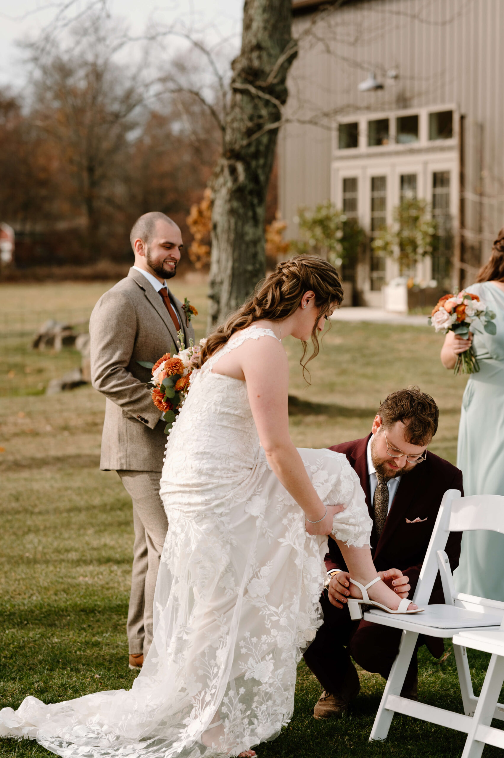 bride's brother helping to adjust the straps on her heels before her wedding ceremony, groom smiling behind them