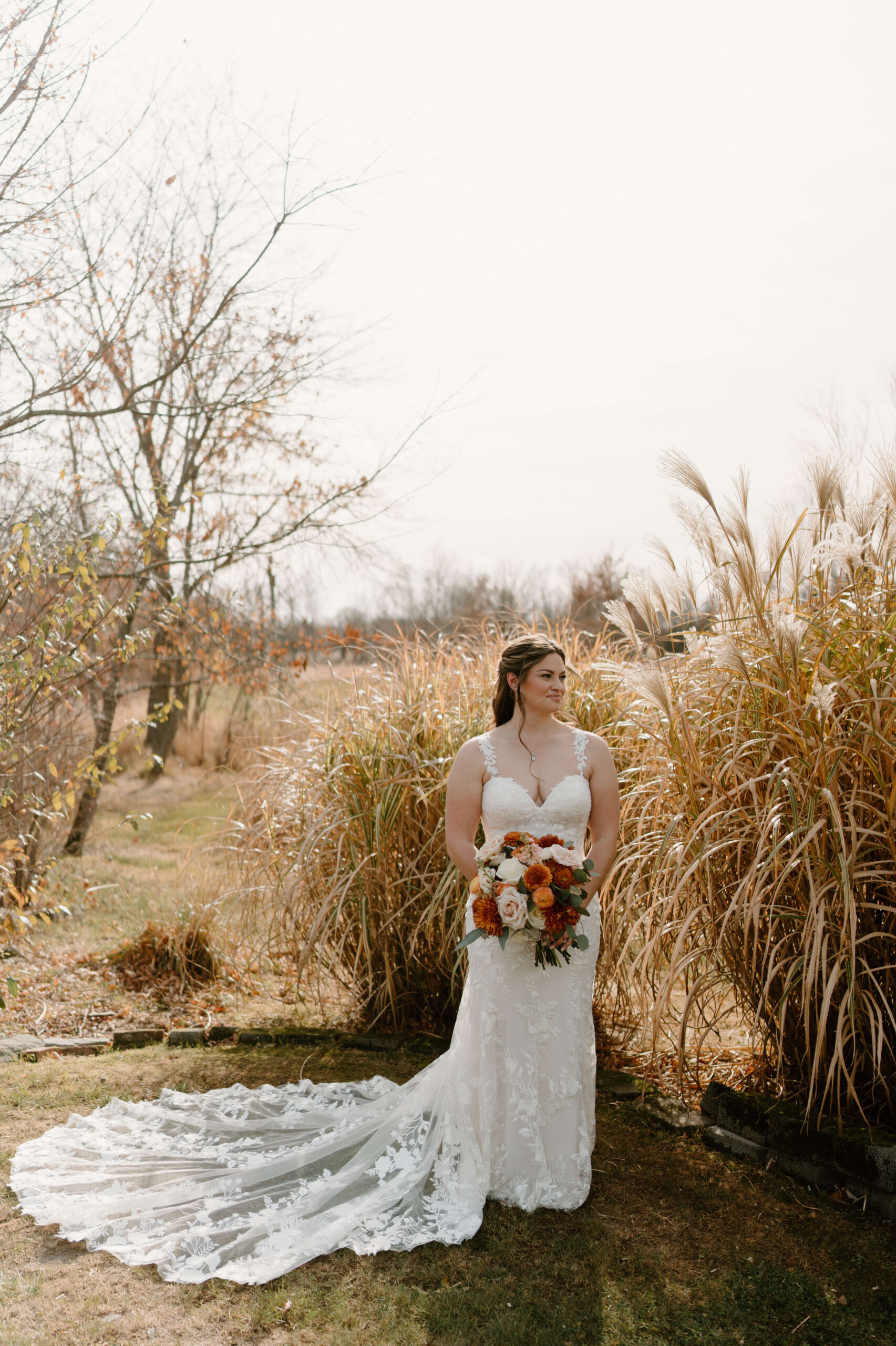 bride in beautiful lace wedding dress with train extending behind her, holding a large bouquet of orange, white, and red flowers, smiling softly off-camera