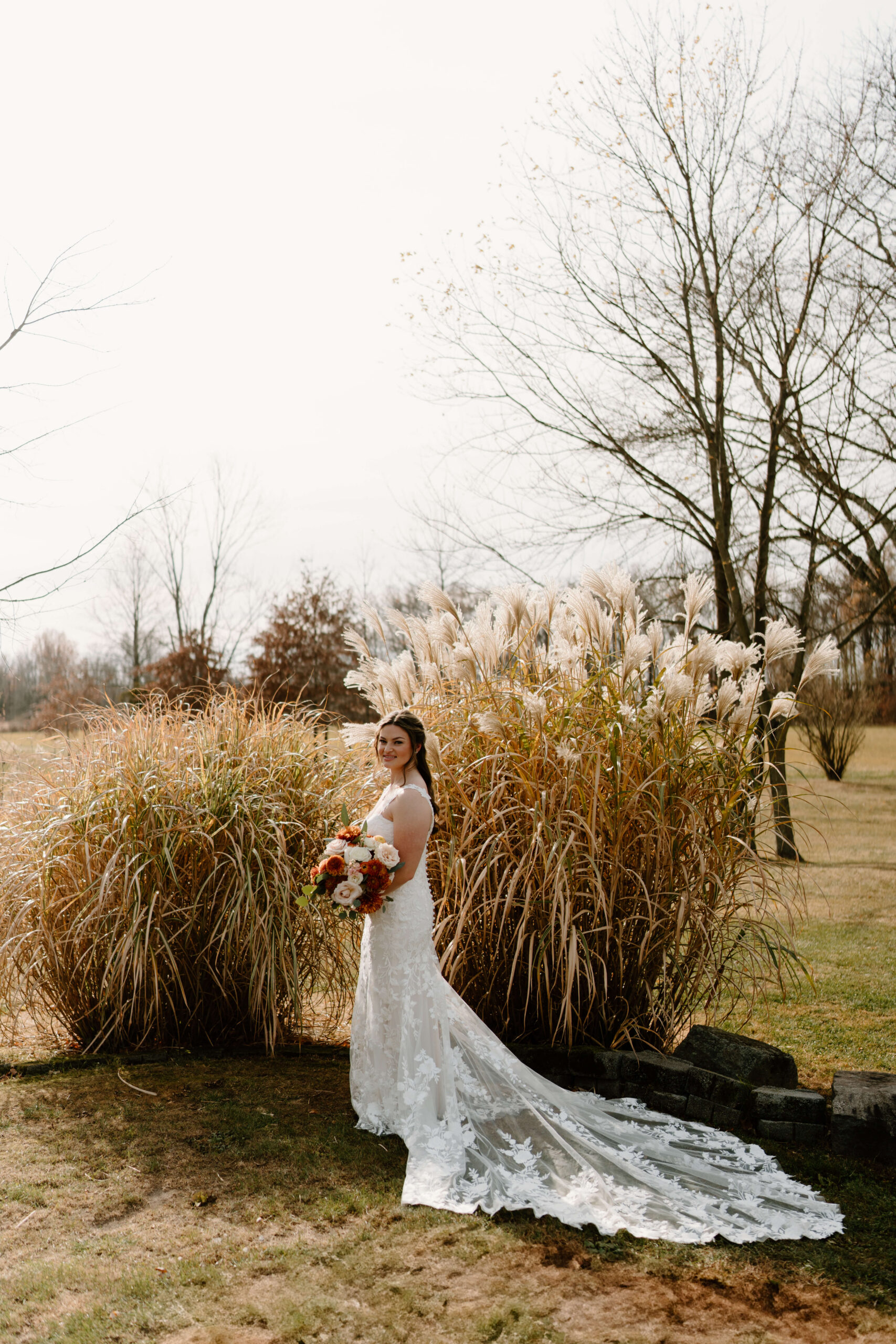 bride in beautiful lace wedding dress with train extending behind her, holding a large bouquet of orange, white, and red flowers, smiling at the camera