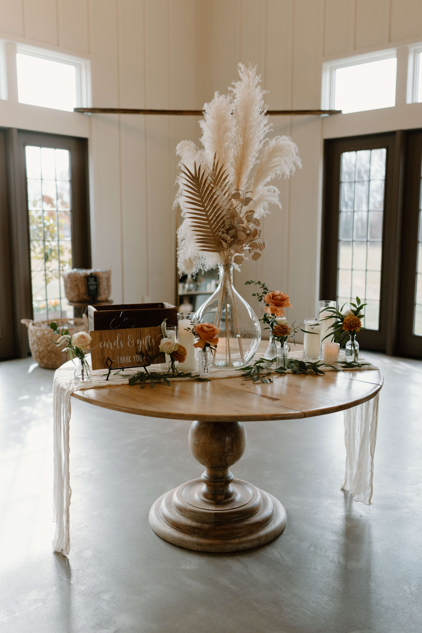 Wedding welcome table in a white walled room, displaying a card box, pampas grass, and other florals