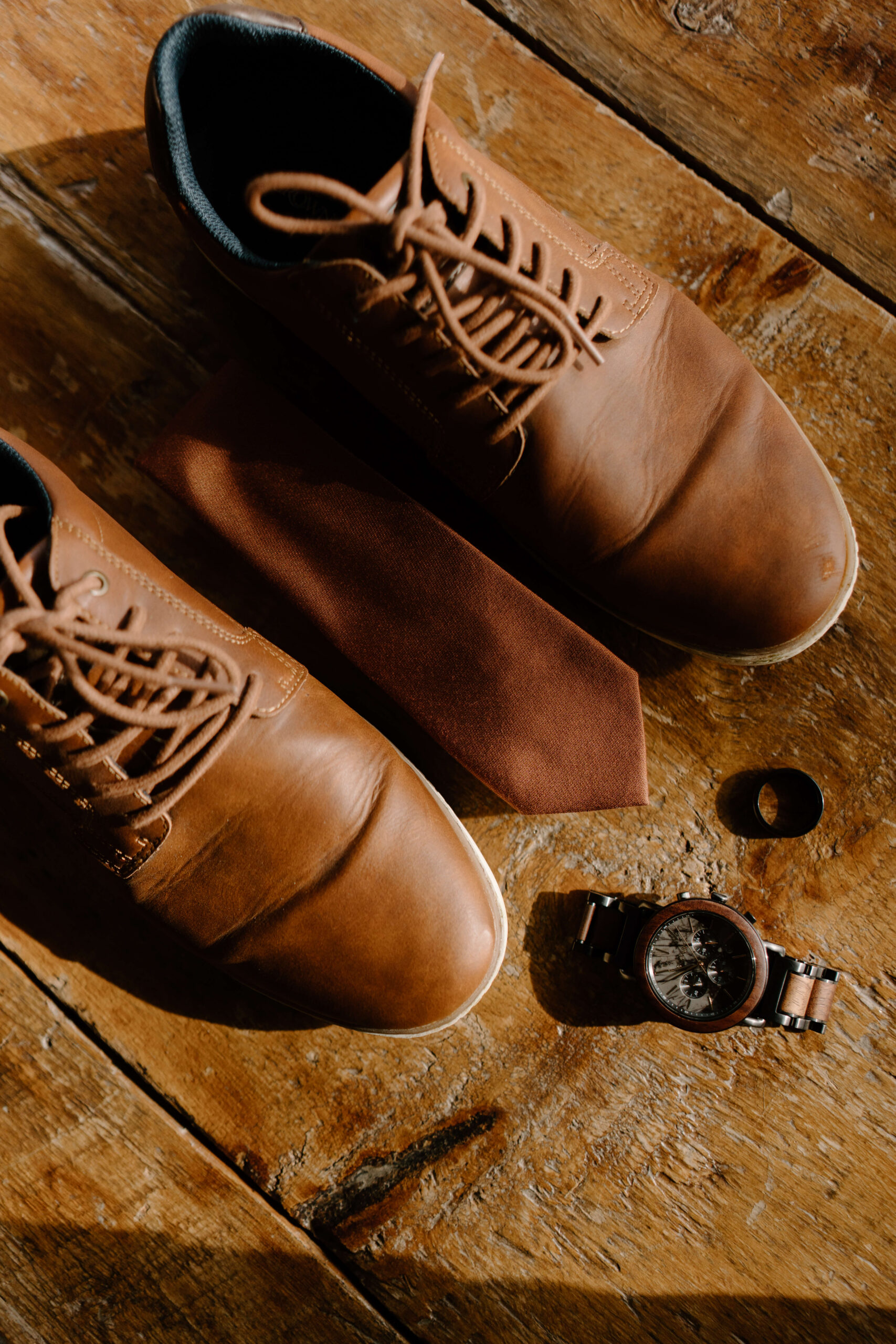 groom's brown dress shoes, watch, tie, and wedding band laid out on the surface of a rustic wooden table