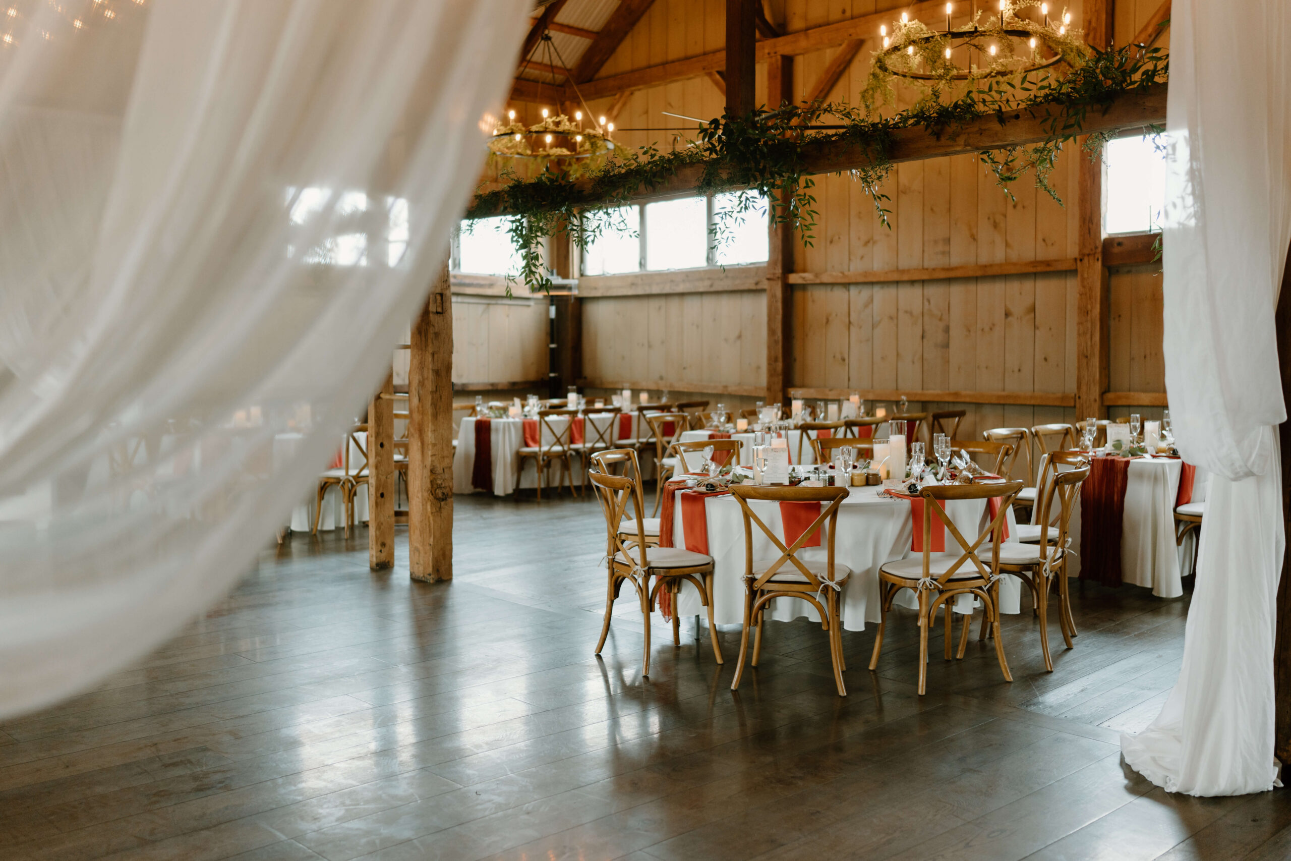 wedding reception space inside a barn, featuring greenery wrapping around the rustic beams, white linen tablecloths and rust-orange linen napkins. 