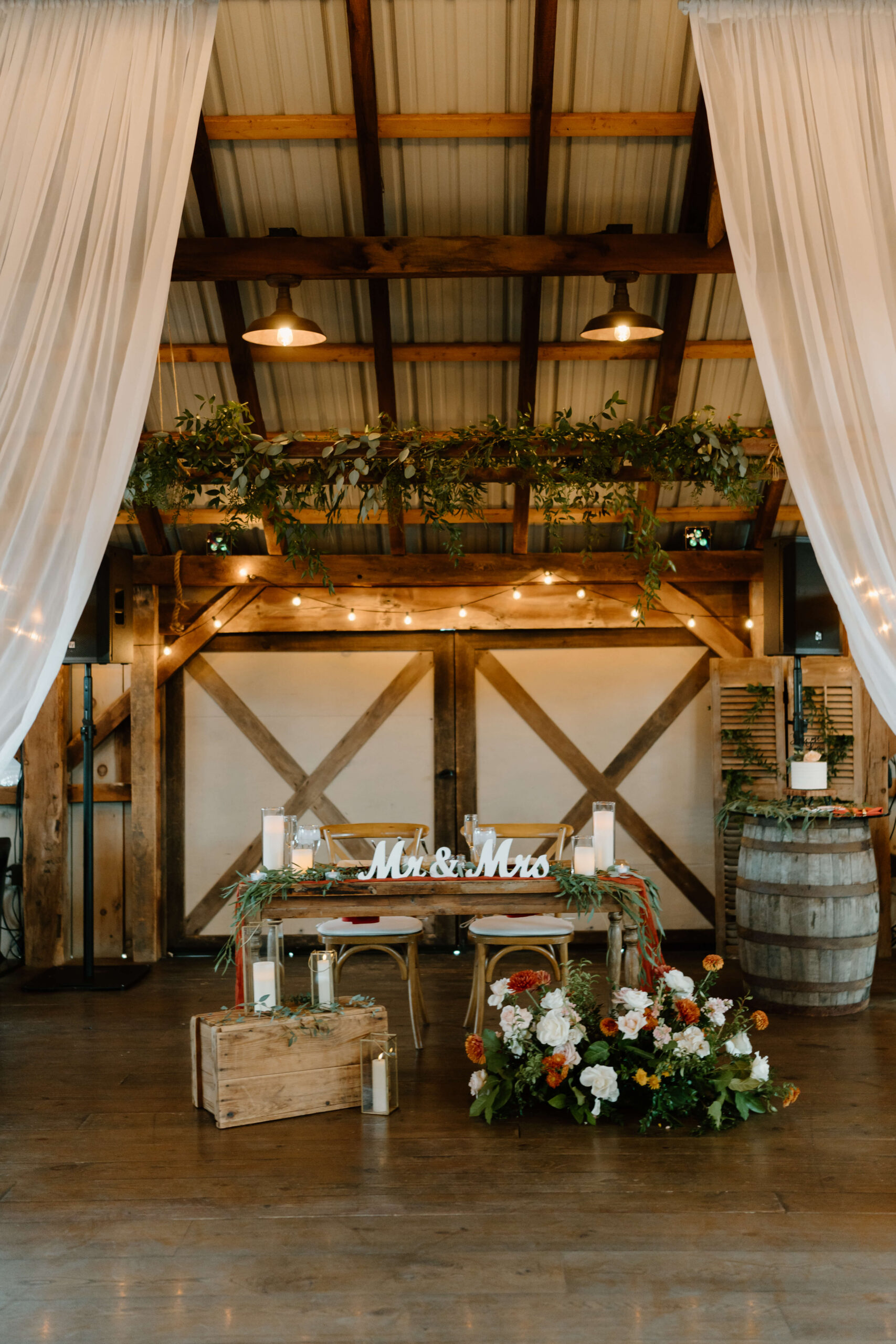 rustic wooden sweetheart table featuring a sign reading "Mr & Mrs", decorated with beautiful orange, white, and greenery floral arrangements and candles in an old barn
