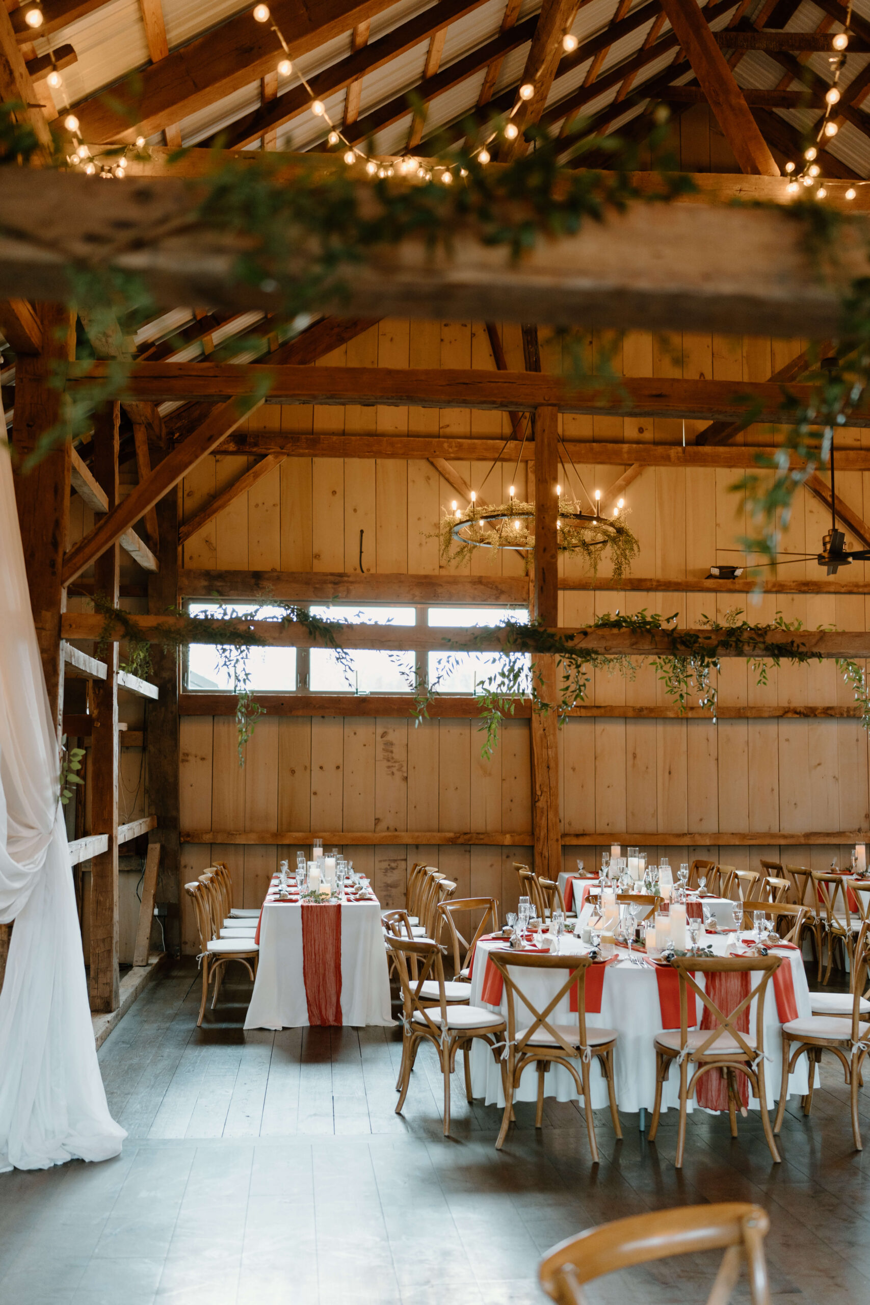 wedding reception space inside a barn, featuring greenery wrapping around the rustic beams, white linen tablecloths and rust-orange linen napkins. 