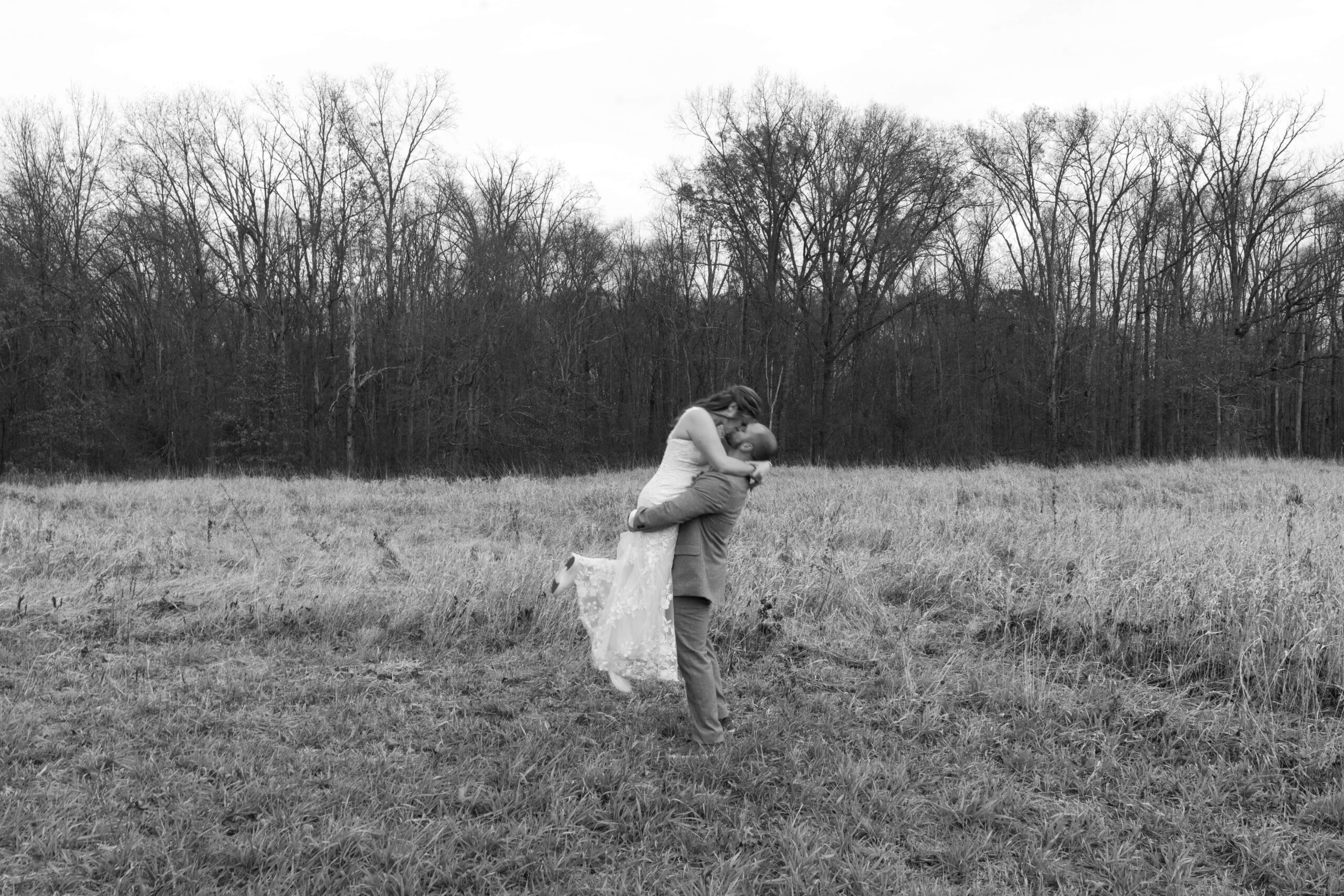black and white image of groom lifting bride and kissing her in a field on wedding day