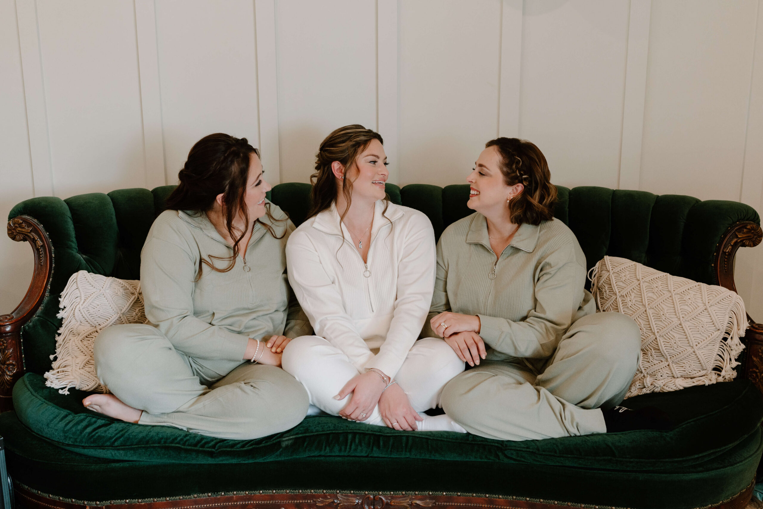 bride (white pajamas) sitting on a green couch with two bridesmaids (green pajamas) smiling at each other