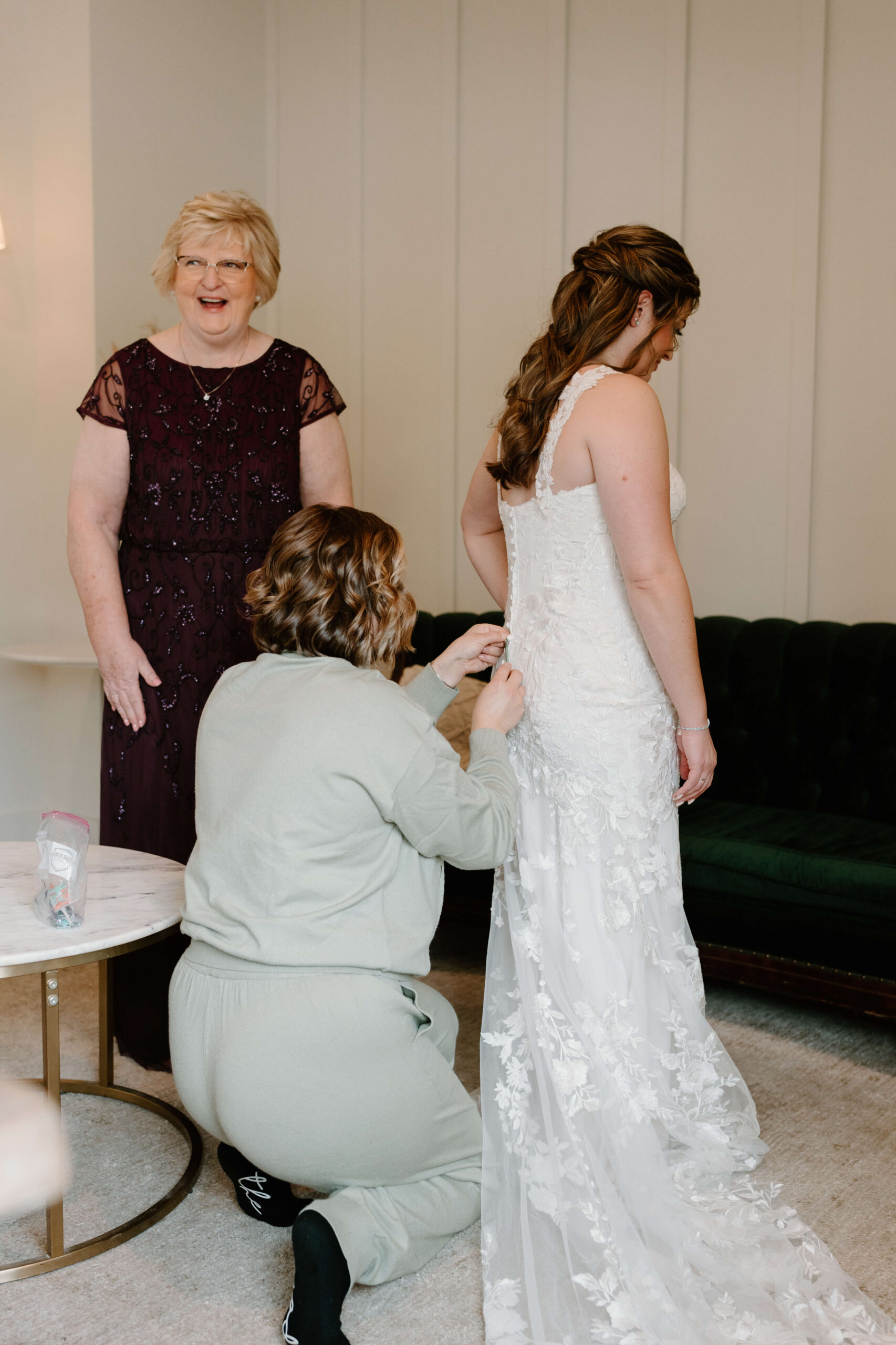 bridesmaid helping to button bride into her wedding dress, while mother of the bride smiles off-camera